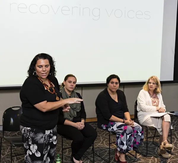 Three women sitting down while another is standing and talking to an unseen crowd.