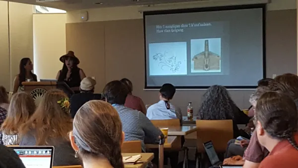 Group of people sitting in front of a presentation given by two Haida individuals.