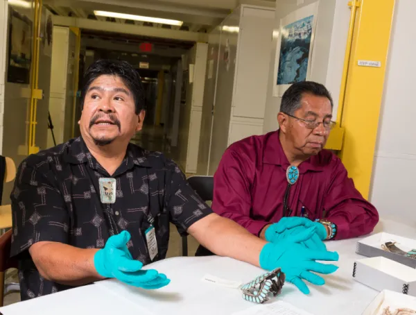 Two men sitting at a table with turquoise jewelry in front of them.
