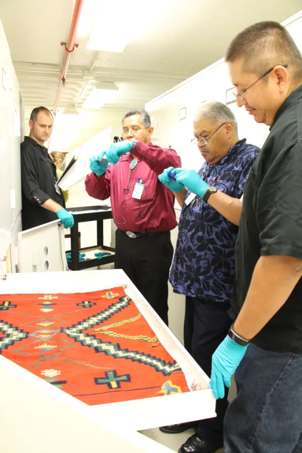 Four men looking at a red Zuni textile with a green geometric pattern.