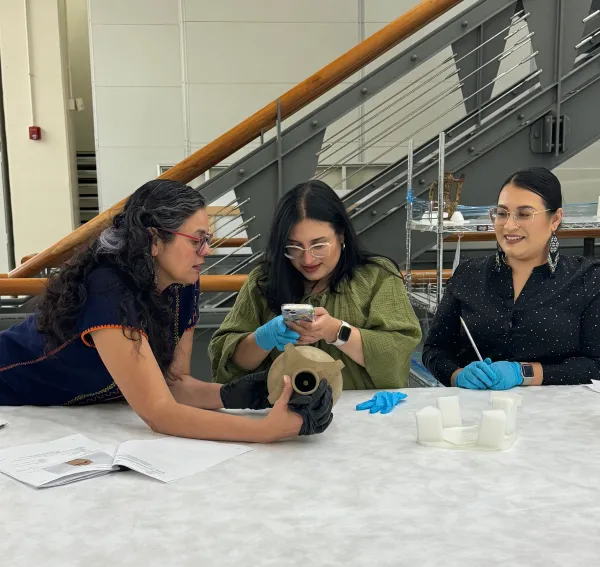Three women looking at a ceramic vessel enclsoed in a rectangular pottery frame. Tania is holding the vessel on its side, with the top opening facing the front, with a small hole seen at the bottom of the vessel.