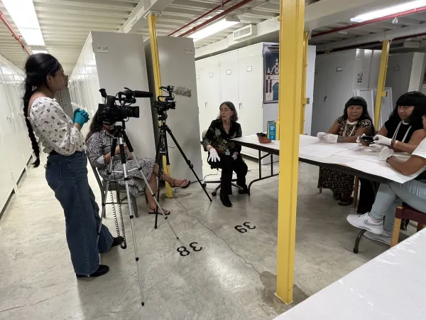 A group of Wauja women sitting around a table while two other women are video and audio recording their conversation.