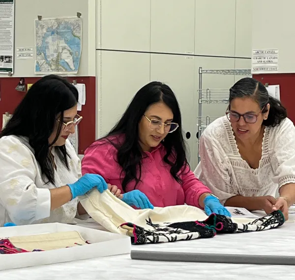 Three woman looking down at a white huanengo (shirt) embroidered with a black floral pattern and green and pink thread around the neckline and arm holes.