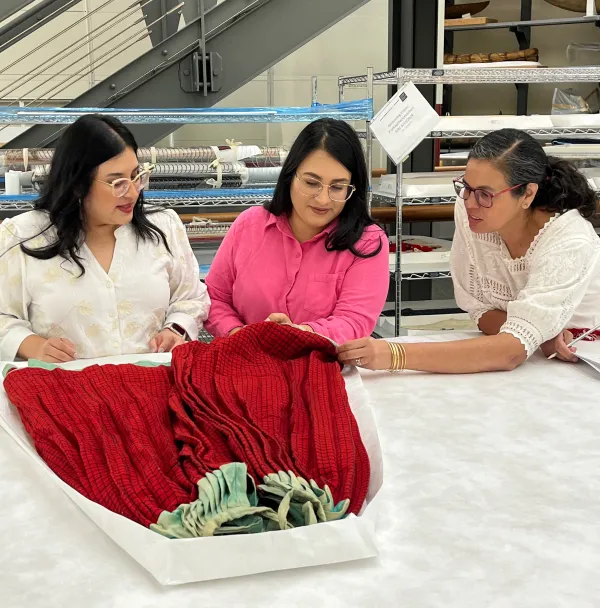 Three women looking at a red skirt with a black checkered pattern and faded green trim around the waistband and lower end of the skirt.