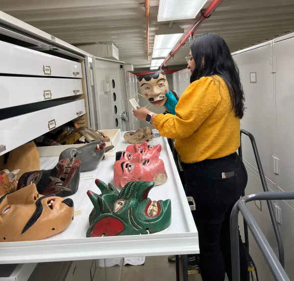 Woman standing on a small ladder, holding a wooden, painted mask in her right hand and looking at the catalog tag. A tray of more painted masks (tan, green, pink, black, and gray) are pulled out in front of her.