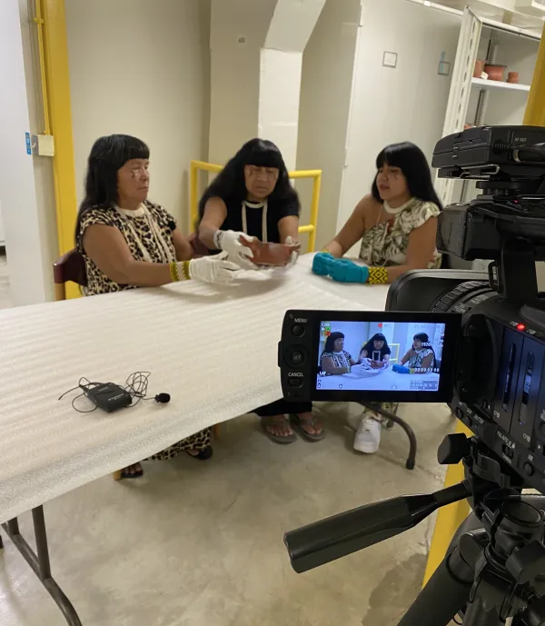 Three women sitting at a table examining a ceramic pot; same image seen through a camera in the foreground.