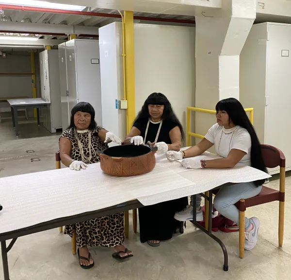 Three woman sitting at a padded table looking at a ceramic vessel.