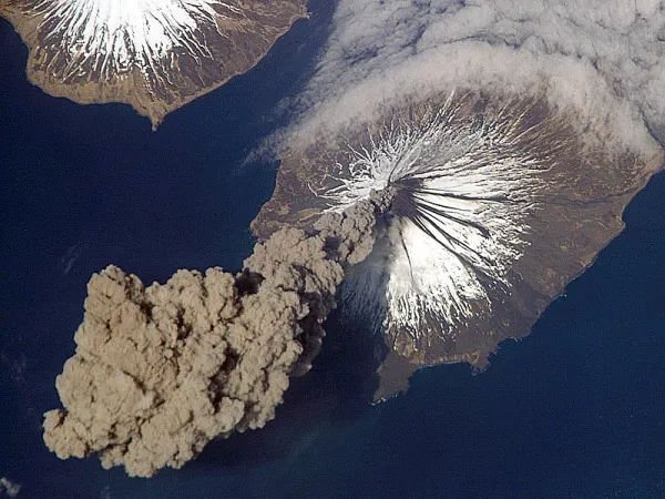 Aerial view of an ash plume rising from Mount Cleveland volcano, which has snow on its upper slopes.
