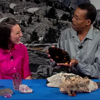 Maggy Benson and geologist Dr. Michael Wise at a table that has gems, minerals, crystals. He is holding a large Quartz gem.