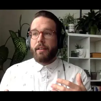 Climate scientist Jeremy Hoffman, a light-skinned, bearded man wearing headphones and white shirt, sitting in front of a bookcase with plants on it