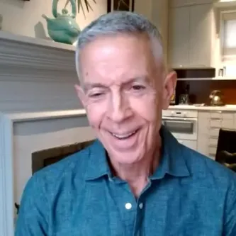 A smiling man with short gray hair wearing a blue shirt. Behind him is a teapot on a shelf and a kitchen counter.