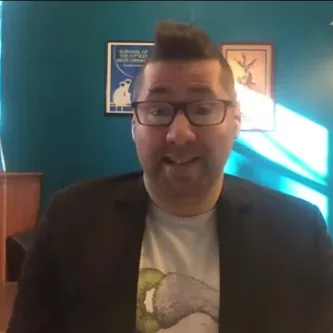 Video screenshot of a light-skinned man with dark hair and glasses, sitting in a room with a blue wall behind him. He is wearing a black jacket over a t-shirt with a design on it.