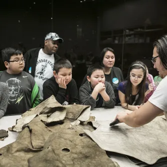 School students around a table, watching a man softening a moose hide with a stone scraper