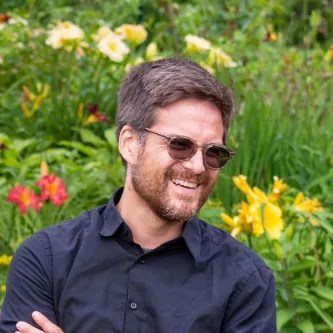 A man with short brown hair, beard, and sunglasses. He is smiling and wearing a navy blue shirt. There are flowers in the background.