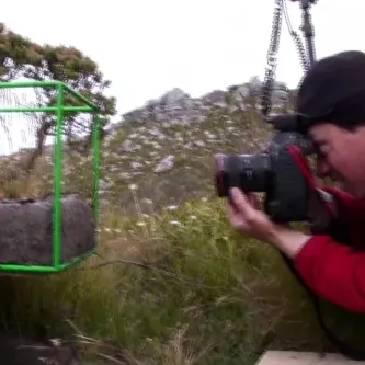 A man in a red jacket holding a camera and squatting to take a photo of a green biocube in the grass.