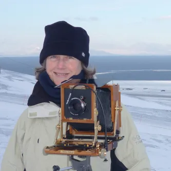 Dornith Doherty, standing in a snow-covered landscape with the ocean in the distance. She has a white face and light brown hair, and is wearing a ski cap and winter coat and is standing behind a camera on a tripod.
