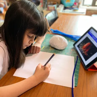 A girl drawing a jellyfish on a sheet of paper with a tablet in front of her.