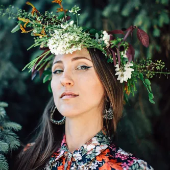 Jenny Kendler wearing a crown of flowers and a floral print shirt. She is a white woman with pale skin and long brown hair