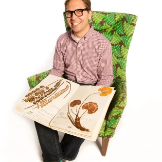 A man sits in a green chair and holds a book of botanical specimens. 