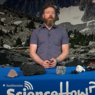 A man with light skin tone and a beard standing behind a table with six different rocks on it.
