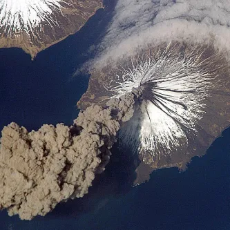 Aerial view of an ash plume rising from Mount Cleveland volcano, which has snow on its upper slopes.