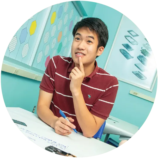 A teenage boy sitting at a table with a pen in his hand. He is touching his chin with one finger, as if he is thinking.