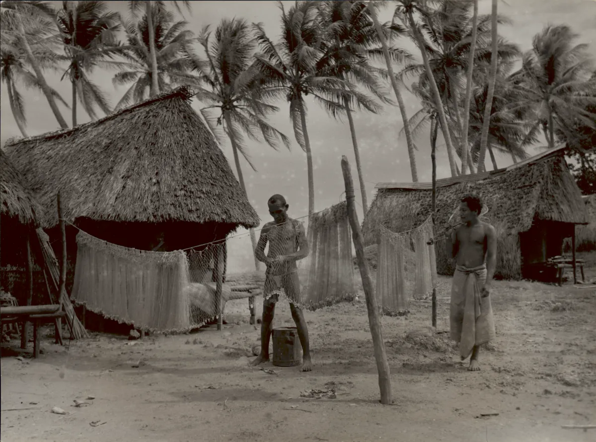 1947 photo of two Trobriand men, one weaving a fish net from pandanus fiber with shells added for weight; two pole and thatch houses in background.