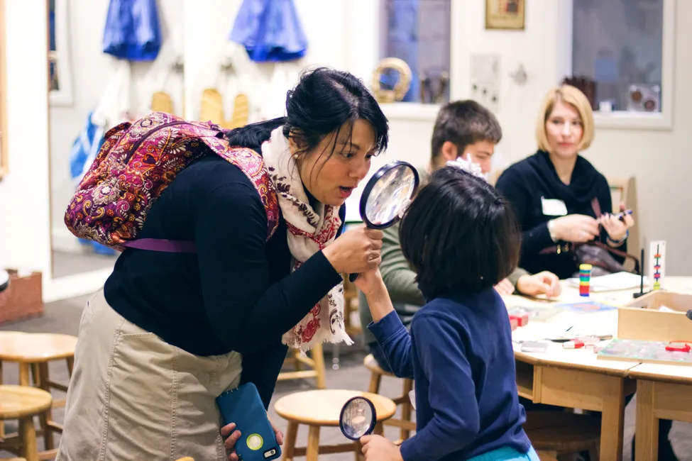 A mother looks at her child through a magnifying glass in a classroom with tables and stools.
