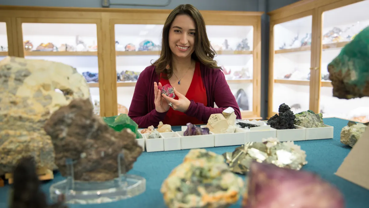 A woman holding a reddish-pink mineral while standing at a table that has many other minerals, and gems on it.