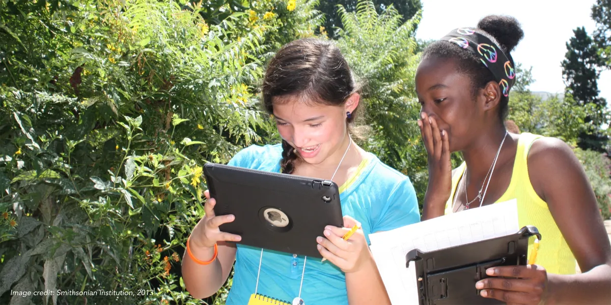 Two girls smiling, looking at a tablet while standing in a garden with tall shrubs.