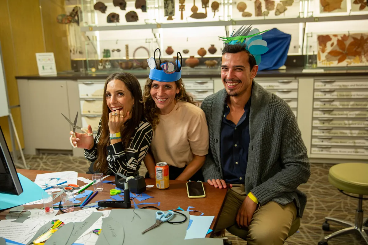 Two women and a man sitting at a table that is covered with craft-making supplies, such as paper, scissors, and markers. The man and one woman are wearing paper hats adorned with hand-drawn fish and a marine reptile.