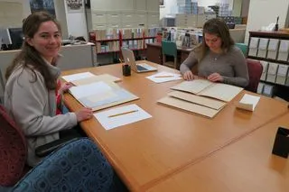 Two women sitting at a desk looking at archival materials.