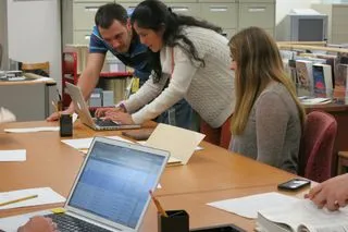 Two people hunched over a computer; one person sitting at a desk.