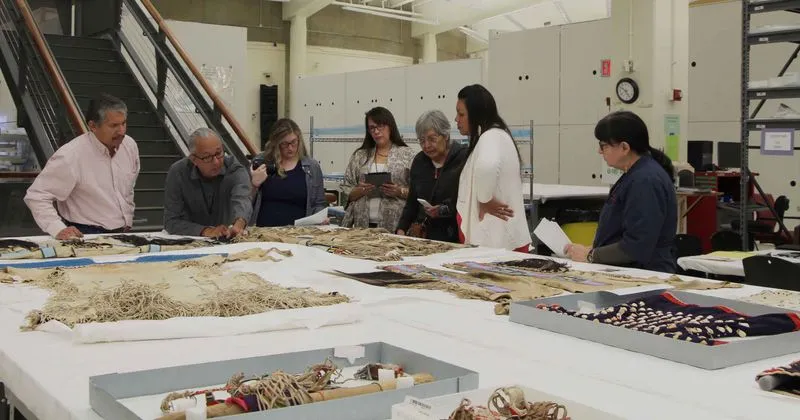 Group of people standing around a table with various Apsáalooke men’s shirts and other buckskin clothing on laid out.