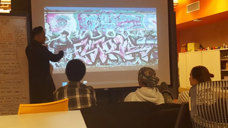Presenter pointing to a Mele mural while students watch.