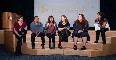People sitting on a steps of a stage in front of a Mother Tongue Film Festival background.