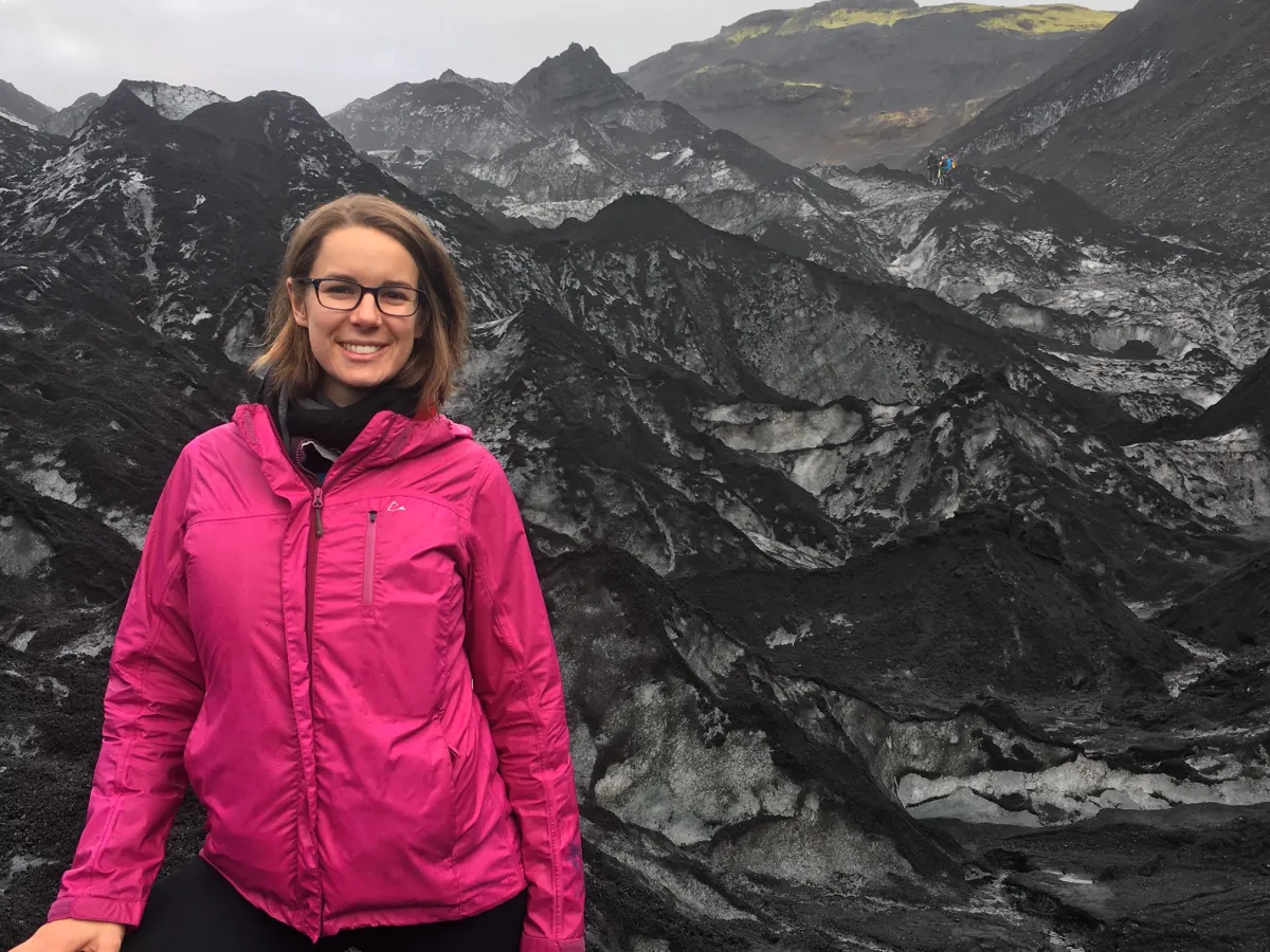 A women in a pink rain jacket standing in front of a rocky, mountainous landscape.