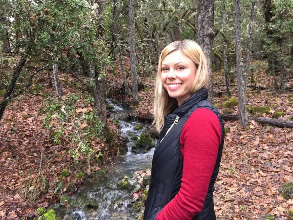 A woman standing by a creek in the woods. Brown leaves cover the ground.