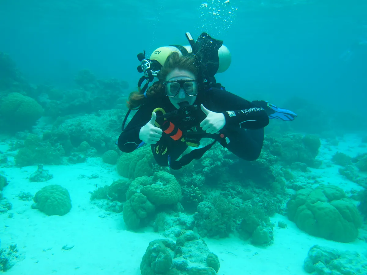 Andie Chan in scuba gear, swimming over the ocean floor giving a two thumbs-up sign.