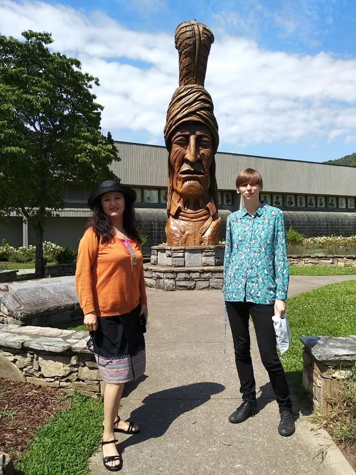Two women stand in front of wood carving outside 