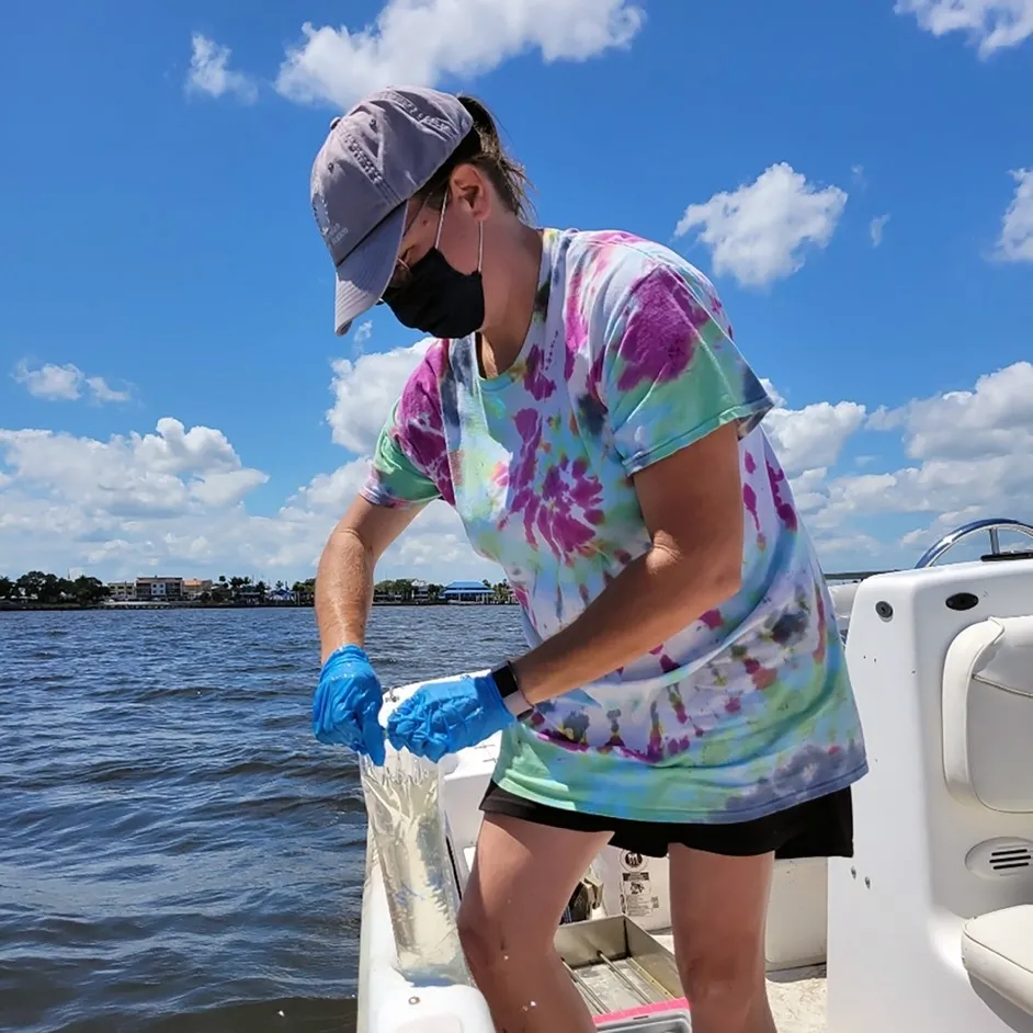 a woman wearing a tie-dyed shirt and blue nitrile gloves and standing on a boat seals a clear plastic bag containing a water sample