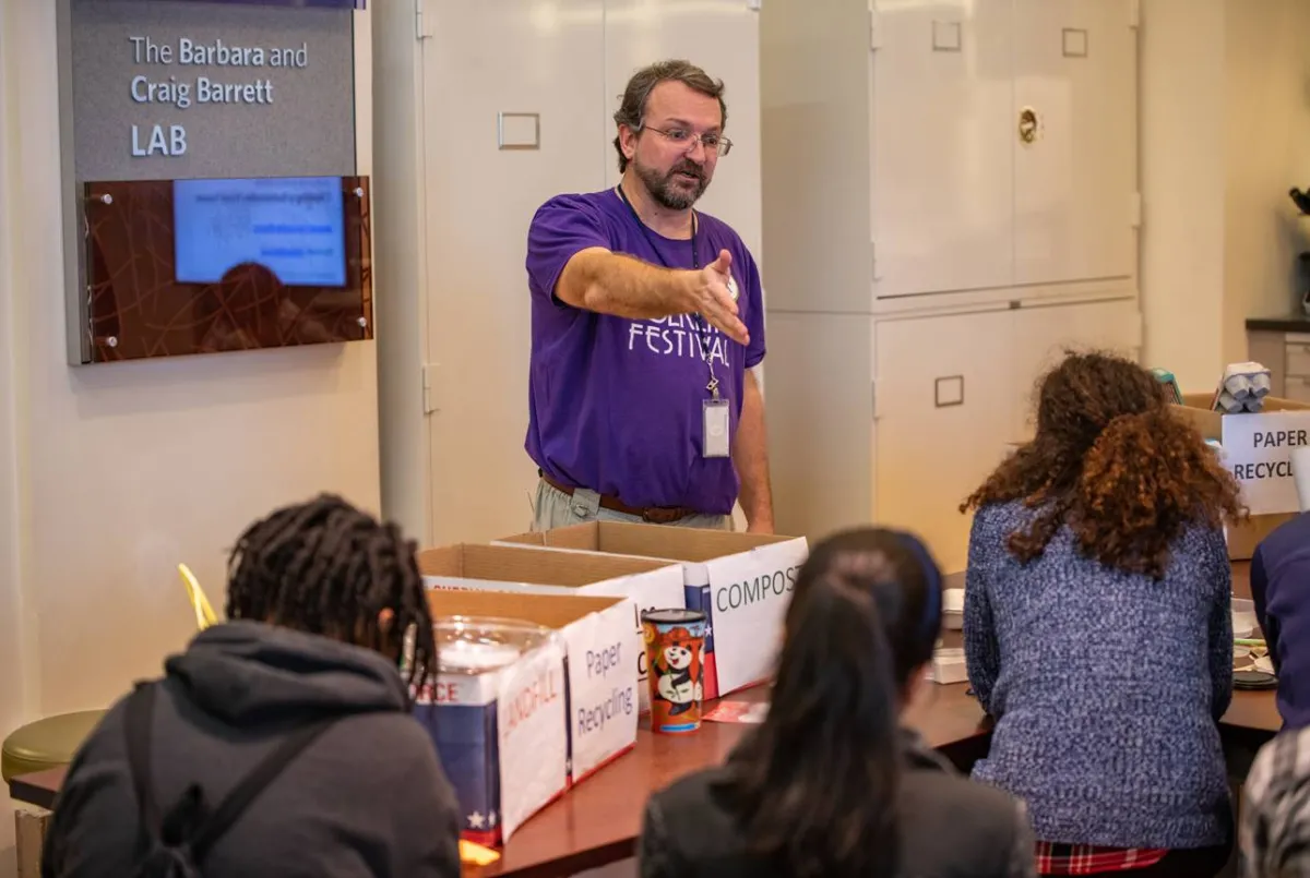Eric Hollinger gestures as he speaks to some teenagers while standing behind a table with sorting boxes for recycling and landfill waste.