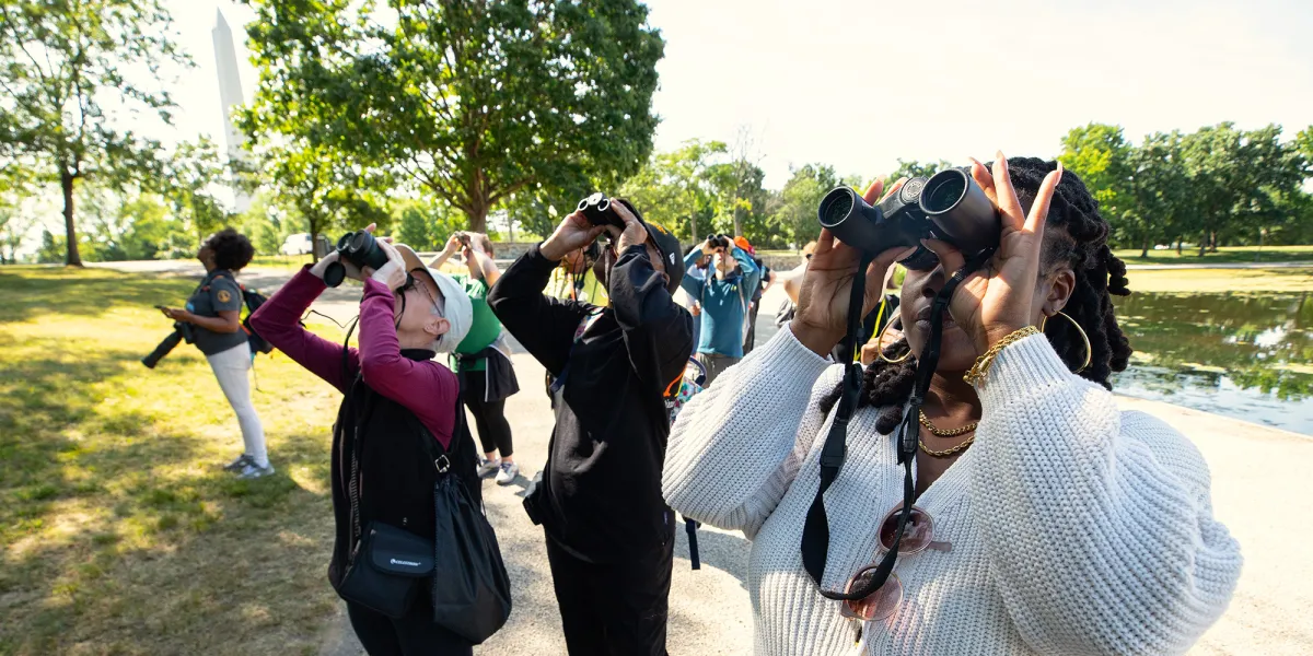 A group of about eight people, including some medium- and light-skinned people, standing on a path on the National Mall and looking up through binoculars. One woman is holding a camera. The Washington Monument is in the background along with some green, leafy trees.