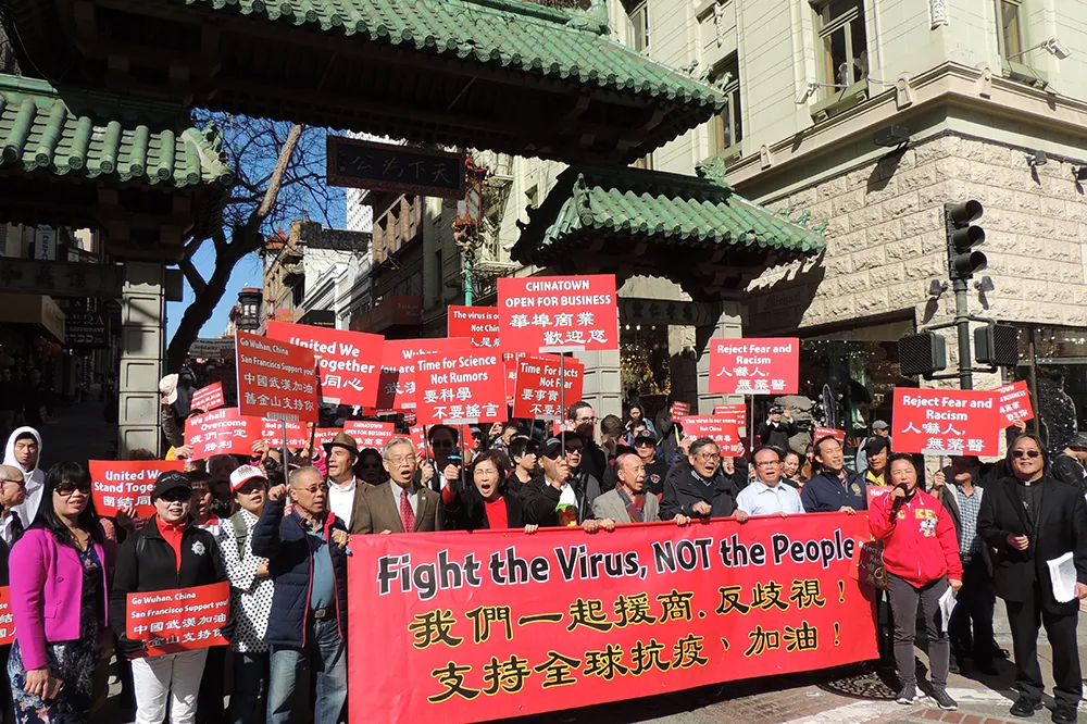 A group of three dozen or more people holding red signs and a large red banner that reads, "Fight the Virus, Not the People."