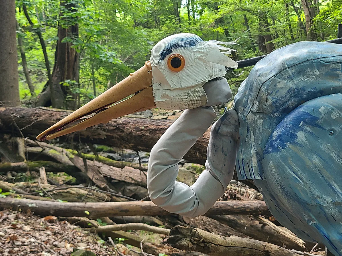 Close-up of the head and upper body of a blue heron puppet in a forest
