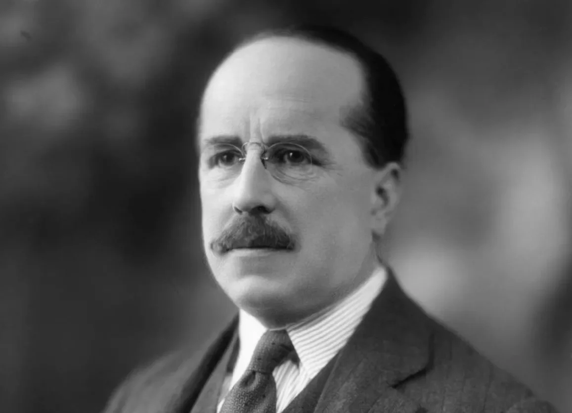 A serious, light-skinned man with glasses and a mustache looks past the camera