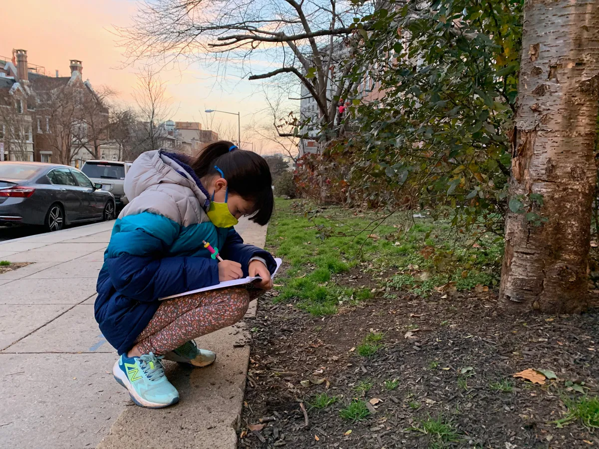 A girl on a city sidewalk, squatting next to a grassy area and taking notes in a notebook.