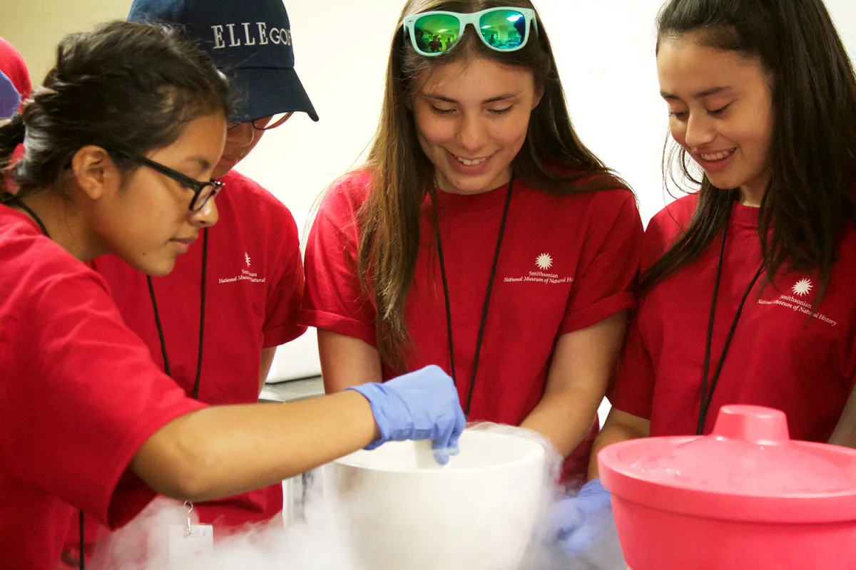 Four teenagers in red t-shirts and blue gloves stand around a white bucket with smoke or steam coming out of it.