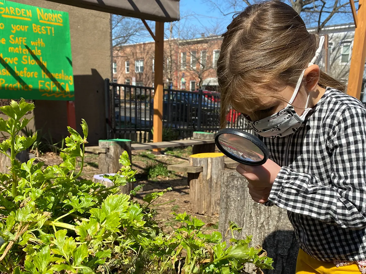 A young girl wearing a mask looks through a magnifying glass at some green plants.