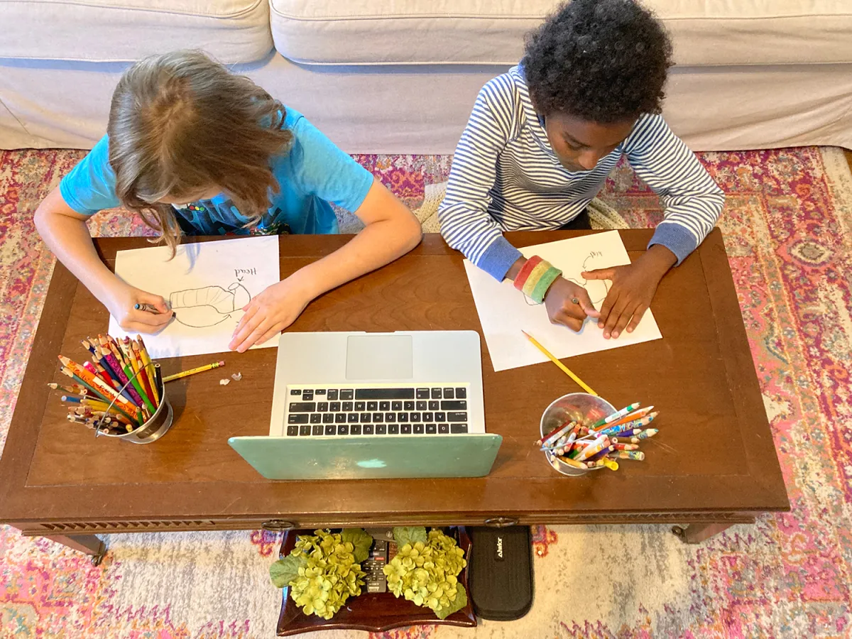 Two children drawing while sitting at a table with a laptop open in front of them